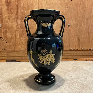 May include: A black ceramic vase with two handles, featuring gold floral patterns on the front and gold trim along the rim and base. The glossy surface reflects light, highlighting the intricate gold botanical designs and the classic amphora shape.