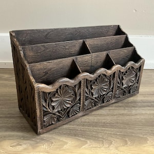 May include: A dark brown wooden desk tidy with four compartments. The front features carved floral designs. The tidy is angled, with a scalloped edge along the bottom. The wood grain is visible.