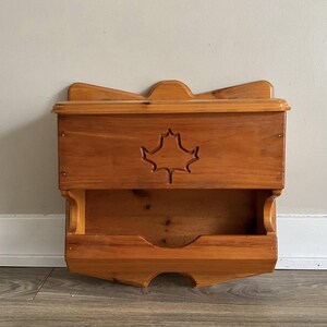 May include: A wooden wall shelf with a carved maple leaf design. The shelf has two compartments, one above the other. The shelf is made of light brown wood and has a rustic appearance.