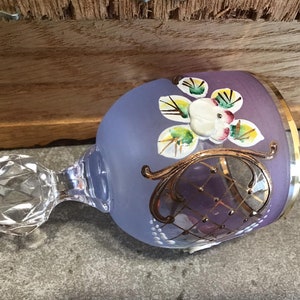 May include: A purple glass goblet with a gold rim and painted floral design. The goblet has a clear glass stem with a faceted knob.
