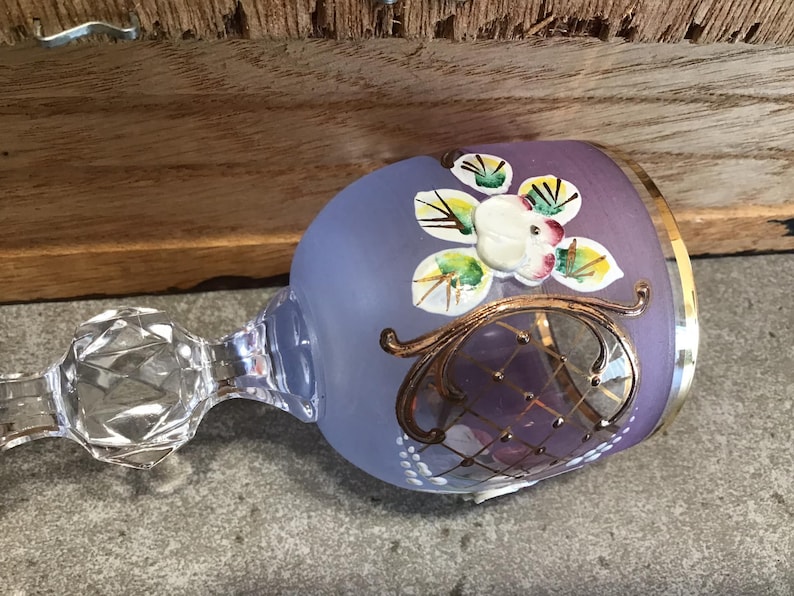 May include: A purple glass goblet with a gold rim and painted floral design. The goblet has a clear glass stem with a faceted knob.