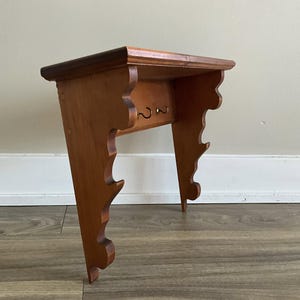 May include: A wooden wall shelf with a decorative, scalloped design and two hooks. The shelf is stained a light brown colour.