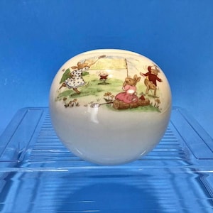 May include: A white ceramic round trinket box with a colorful illustration of rabbits playing in a field. The box has a lid and is decorated with a whimsical scene of rabbits playing with a ball and a flag.