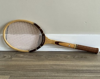 Könnte beinhalten: Vintage Tennis-Schläger aus Holz mit hellbraunem Rahmen, weißen Saiten und einem dunkelbraunen Griff. Der Schläger hat ein braunes Band um den Kopf und das Wort "CHAMPIONSHIP" ist sichtbar. Der Griff ist braun mit einem Dunlop-Logo.