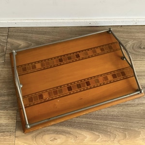 May include: A rectangular wooden serving tray featuring a light brown finish with two parallel decorative inlay bands of darker wood squares. The tray is equipped with a curved, silver-toned metal handle frame that spans the two shorter sides.