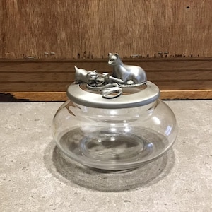 Puede incluir: Un tarro de vidrio transparente con una tapa de plata que presenta dos gatos. La tapa está decorada con un gato de plata sentado sobre una bola de plata, y un gato de plata más pequeño está mirando al gato más grande.