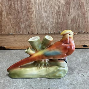 Puede incluir: Una figurita de pájaro de cerámica con un cuerpo rojo, naranja y azul, posada en una rama verde con dos aberturas para flores. El pájaro tiene una cabeza y un pico amarillos.