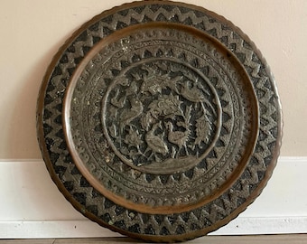 May include: A large, round, copper tray with a decorative design of birds and flowers. The tray has a dark brown patina and a scalloped edge.