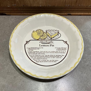 Peut inclure: Un plat à tarte en céramique blanche avec un bord festonné jaune. Le centre présente une recette de tarte au citron avec des illustrations de citrons et une part de tarte. Le texte indique "Rawleigh's Lemon Pie".
