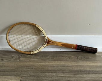 May include: Vintage wooden tennis racket with a light brown frame and a white grip. The racket has a natural wood finish with a dark brown handle and a red accent. The strings are white and the racket is resting on a wooden surface.