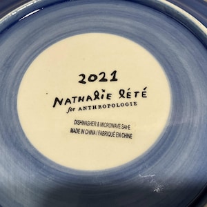 Może przedstawiać: Ceramiczny talerz z niebiesko-białym wzorem. Środek talerza jest biały z napisem "2021 NATHALIE R&Eacute;T&Eacute; for ANTHROPOLOGIE". Talerz nadaje się do mycia w zmywarce i kuchence mikrofalowej. Wyprodukowano w Chinach.
