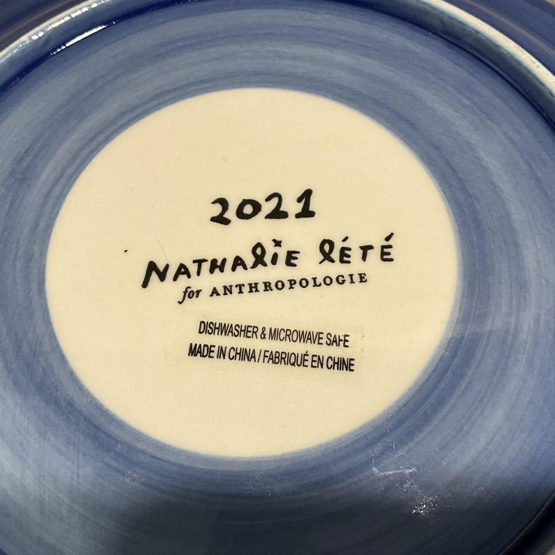 Może przedstawiać: Ceramiczny talerz z niebiesko-białym wzorem. Środek talerza jest biały z napisem "2021 NATHALIE R&Eacute;T&Eacute; for ANTHROPOLOGIE". Talerz nadaje się do mycia w zmywarce i kuchence mikrofalowej. Wyprodukowano w Chinach.