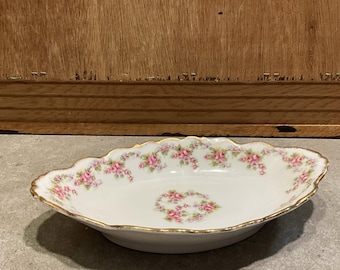 May include: A white, oval-shaped porcelain dish with a scalloped edge. The dish is decorated with pink roses and gold trim. The roses are arranged around the rim and in the center of the dish. A vintage serving piece.