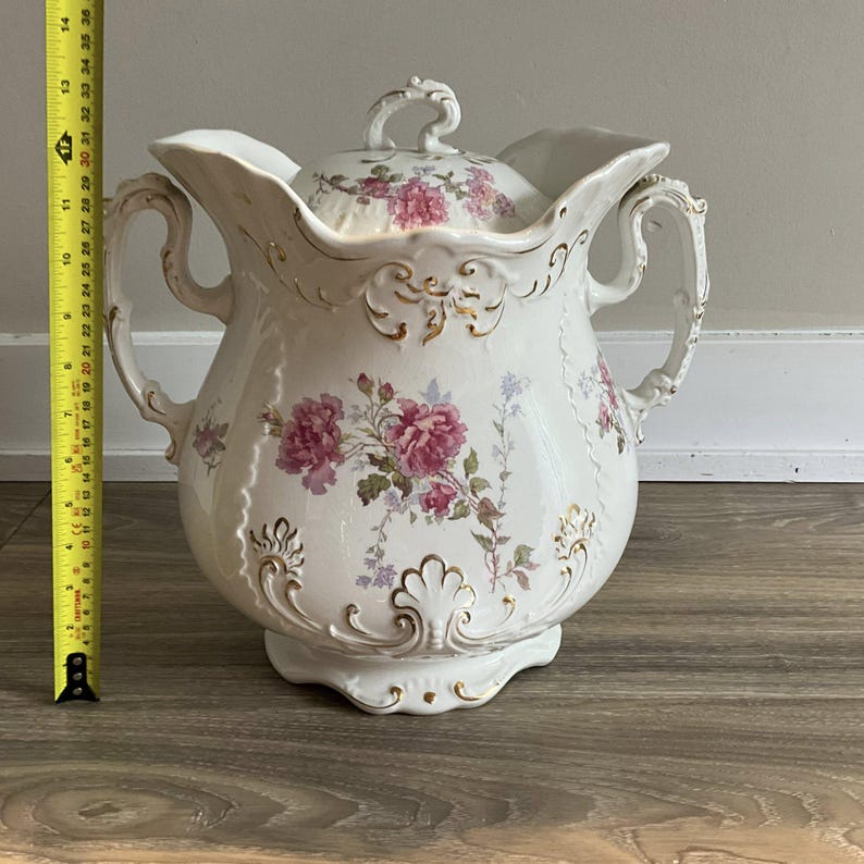 May include: A white porcelain pitcher with a lid, adorned with pink floral patterns and gold accents. The pitcher has two handles and measures approximately 14 inches tall.