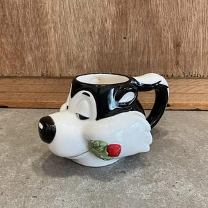 May include: A white ceramic mug with a black and white cartoon of a dog with a red rose on its cheek. The mug has a black handle.