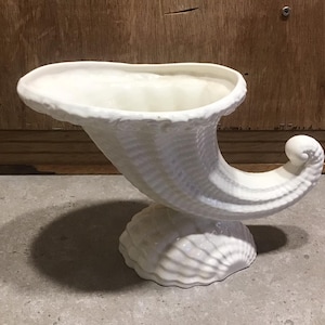 Puede incluir: Jarrón de cerámica blanca en forma de cornucopia con una superficie texturizada. El jarrón tiene forma de cuerno y una abertura ancha en la parte superior. Es una pieza decorativa que se puede utilizar para guardar flores u otros objetos pequeños.