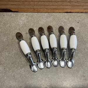 May include: Six white ceramic cabinet knobs with a chrome finish. The knobs have a rounded oval shape with a slight curve at the top.