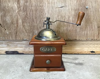Puede incluir: Un molinillo de café de madera con una parte superior de latón y una manivela. El molinillo tiene un cajón con una perilla de latón y la palabra "Cafe" escrita en la parte delantera.