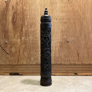 May include: A dark brown, cylindrical wooden object with intricate carvings of animals, possibly elephants and dragons. The top has a decorative finial, and the base is slightly wider. The object is standing upright against a wooden background.