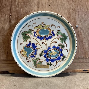 Może przedstawiać: Biały ceramiczny talerz z niebieskim i zielonym wzorem kwiatowym. Talerz ma czarną kropkowaną obwódkę i złotą etykietę z napisem "Hand Painted, Honiton Pottery, Devon, England".