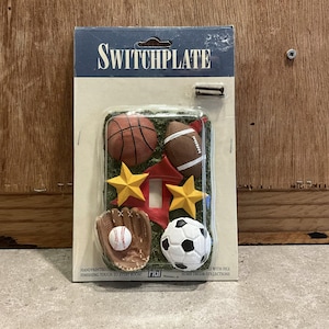 Pode incluir: Uma placa de interruptor de luz com tema desportivo com uma bola de beisebol, uma bola de basquete, uma bola de futebol americano, uma bola de futebol e duas estrelas. A placa é pintada à mão e diz "Switchplate" na parte superior e "Trigi Home Decor Collections" na parte inferior.