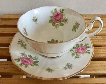May include: White teacup and saucer set with pink rose floral pattern and gold trim. The teacup has a curved handle and a scalloped rim. The saucer also has a scalloped edge. The set is on a wooden surface.