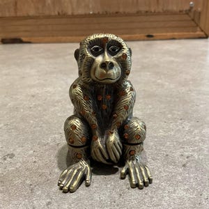 May include: A bronze-colored monkey figurine with orange crystal accents. The monkey is in a seated position with its hands resting on its knees. The figurine has detailed fur and facial features, and is likely a decorative item.