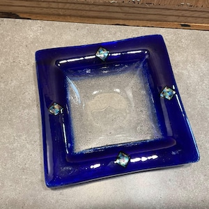 May include: A square glass dish featuring a deep cobalt blue border and a clear, textured center. Four small, square iridescent glass accents are embedded into the blue rim, positioned at the center of each side of the square dish.