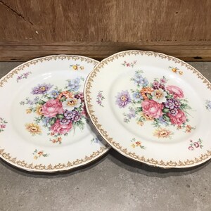 May include: Two white ceramic plates with a gold rim and a floral design. The flowers are pink, purple, yellow, and white.