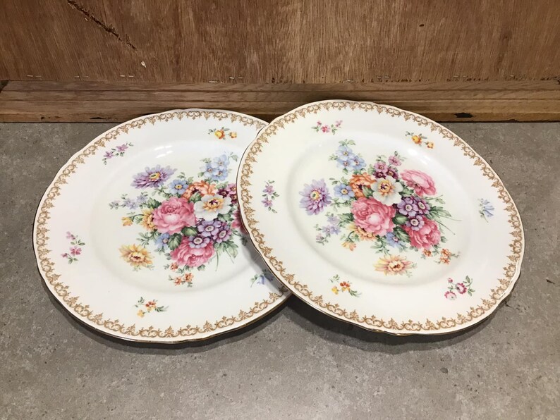 May include: Two white ceramic plates with a gold rim and a floral design. The flowers are pink, purple, yellow, and white.