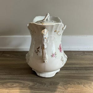 May include: A white porcelain pitcher with a lid, featuring floral and gold accents. The pitcher has a curved handle and a decorative spout. The floral design includes pink and purple flowers. The pitcher is a vintage style.