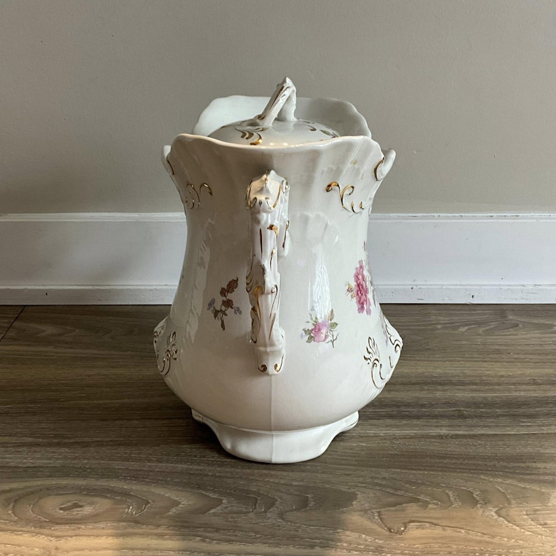 May include: A white porcelain pitcher with a lid, featuring floral and gold accents. The pitcher has a curved handle and a decorative spout. The floral design includes pink and purple flowers. The pitcher is a vintage style.