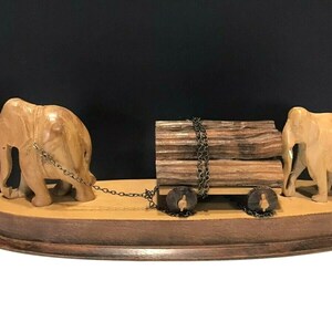 May include: A wooden sculpture of two elephants pulling a cart with logs. The elephants are carved in detail and the cart has wheels. The sculpture is on a wooden base.