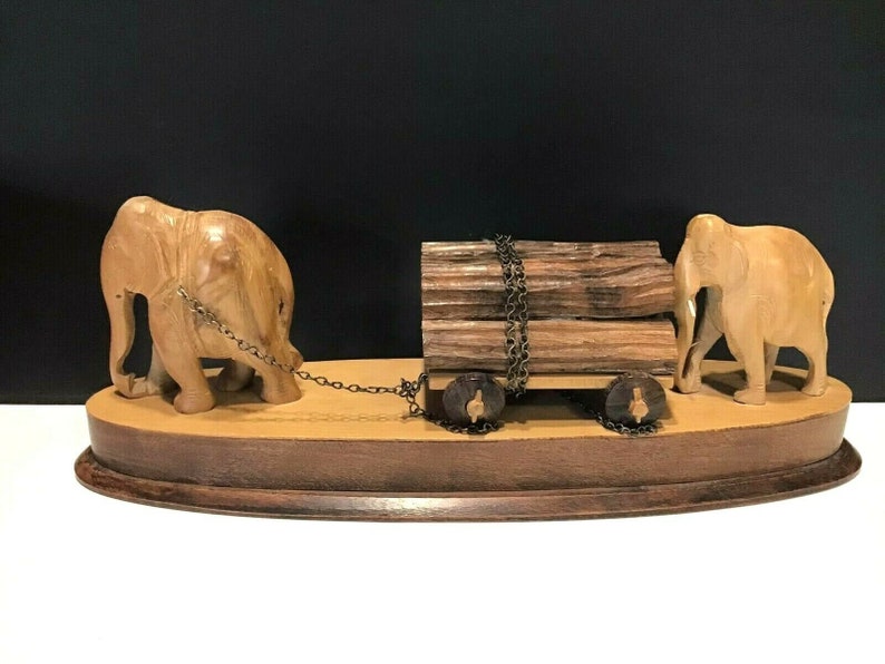 May include: A wooden sculpture of two elephants pulling a cart with logs. The elephants are carved in detail and the cart has wheels. The sculpture is on a wooden base.