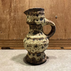 Puede incluir: Una jarra de cerámica vintage con una superficie texturizada. La jarra tiene un asa y un pico. Los colores son una mezcla de marrón, beige y azul. La jarra está sobre una superficie gris.