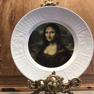 Op de afbeelding: Witte keramische plaat met een reliëfdesign rond de rand. Het midden van de plaat heeft een gedrukt beeld van de Mona Lisa.