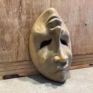 May include: A carved wooden mask with a neutral expression. The mask is light brown with visible wood grain and features a prominent nose, closed mouth, and eye openings. The mask is set against a wooden background.