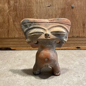 Pode incluir: Pequena estatueta de terracota com um rosto sorridente e braços levantados. A figura tem olhos fechados, um nariz pequeno e uma barba. O corpo é de um tom castanho mais claro e a peça tem um aspeto rústico e feito à mão.
