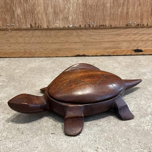 Puede incluir: Caja de madera artesanal con forma de tortuga, de color marrón oscuro. La tapa tiene bisagras y la concha tiene detalles tallados. La tortuga tiene cuatro patas y una pequeña cola.