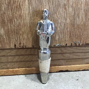 Può includere: Un tappo per bottiglia color argento con una figura in abito che tiene in mano un piccolo oggetto. Il tappo ha una base in sughero ed è progettato per adattarsi a una bottiglia. Lo sfondo è una superficie di legno.