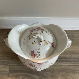 May include: A white porcelain chamber pot with a lid, featuring a floral design in pink and gold. The pot has a scalloped rim and handles, with a matching lid and ornate handle. A vintage decorative item.