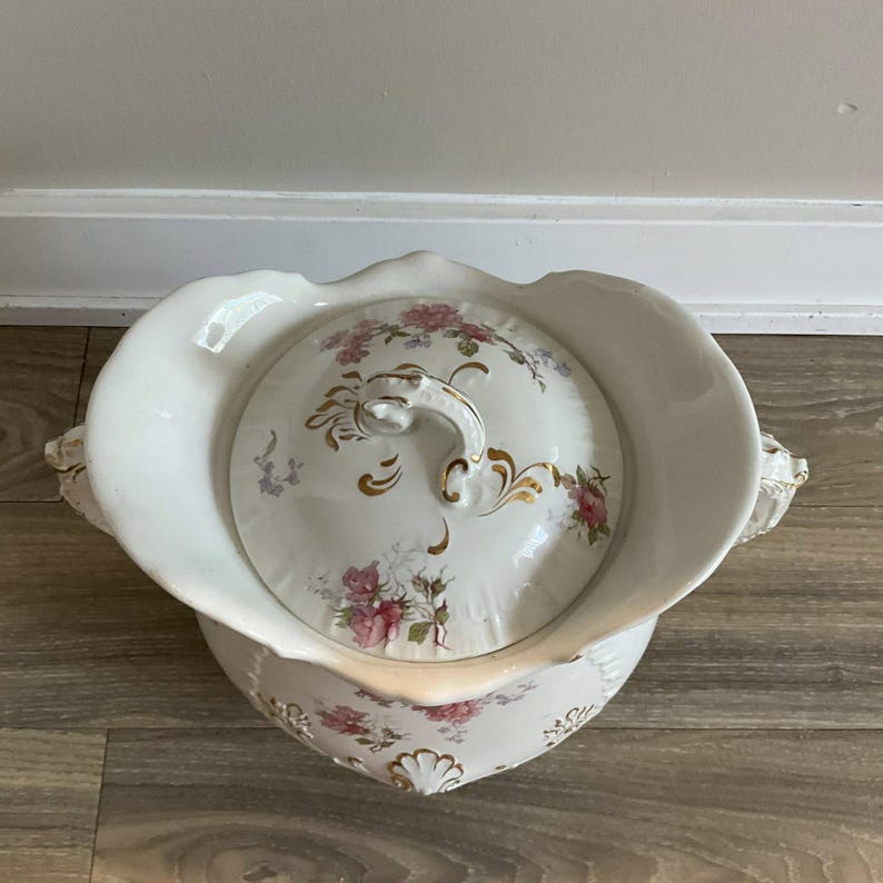 May include: A white porcelain chamber pot with a lid, featuring a floral design in pink and gold. The pot has a scalloped rim and handles, with a matching lid and ornate handle. A vintage decorative item.