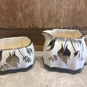 Könnte beinhalten: Ein weißes Keramik-Sahne- und Zuckergefäß-Set mit silbernen Blattdesigns. Das Set verfügt über ein stilisiertes Blattmotiv.