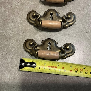 May include: Three antique brass drawer pulls with keyhole details and light brown wooden handles. A Craftsman tape measure shows the pulls are approximately 4 inches long.