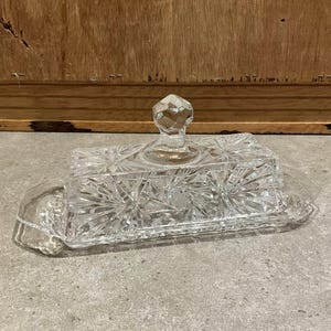 May include: A clear, rectangular glass butter dish with a faceted knob on the lid. The dish features a starburst pattern and small handles on each end. The butter dish is sitting on a gray surface.