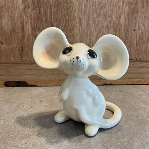 May include: A white ceramic figurine of a mouse with large ears and black eyes. The mouse is sitting with its tail curled around its body.