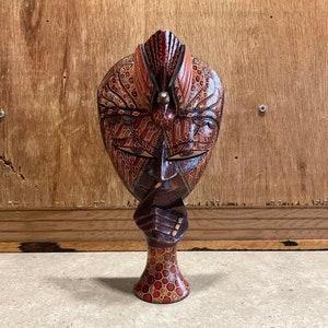 Könnte beinhalten: Eine Holzmaske mit komplizierten roten und braunen Mustern. Die Maske ist in Form eines menschlichen Gesichts geschnitzt und hat einen Vogel auf dem Kopf.