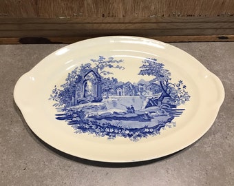 Op de afbeelding: Een witte ovale serveerschaal met een blauw en wit landschapsontwerp. Het ontwerp toont een rivier, bomen en een stenen gebouw.