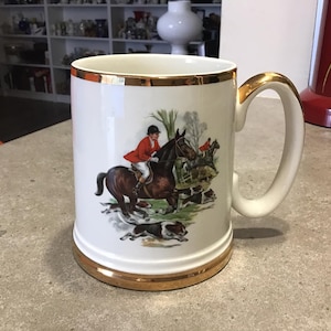 May include: White ceramic beer stein with a gold rim and handle. The stein features a colourful illustration of a person riding a horse with dogs running alongside.
