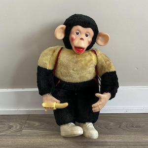Puede incluir: Un juguete de mono de peluche vintage con pelaje negro, pecho amarillo y marrón y tirantes rojos. El mono lleva pantalones negros y zapatos blancos. El mono sostiene un plátano amarillo en su mano.